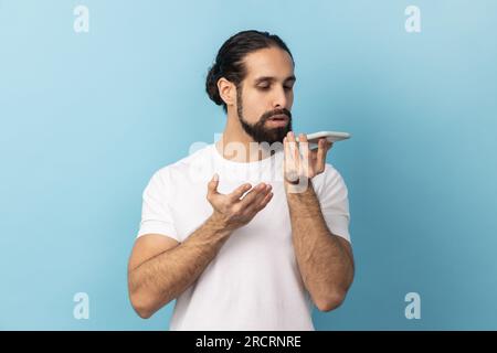Tecnologia Smart Voice. Ritratto di un uomo con la barba che indossa una T-shirt bianca che parla con il cellulare utilizzando l'assistente virtuale e l'applicazione di altoparlanti digitali. Riprese in studio in interni isolate su sfondo blu. Foto Stock