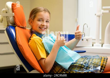 Studentessa di buon umore si mostra alzata con il pollice mentre si siede in poltrona dopo un appuntamento regolare con il dentista. Ragazza su appuntamento dentistico. Controllo regolare Foto Stock