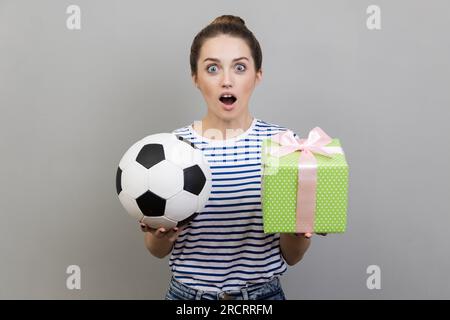 Ritratto di una donna sorpresa che indossa una T-shirt a righe che guarda la fotocamera con gli occhi grandi, che tiene la palla da calcio e la scatola regalo verde. Riprese in studio in interni isolate su sfondo grigio. Foto Stock