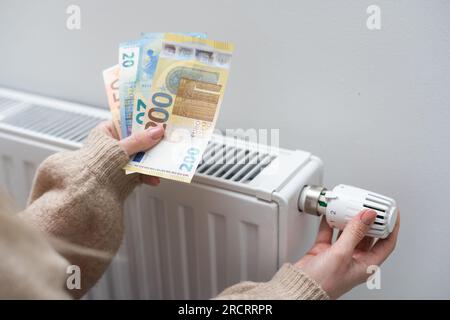 Donna che tiene contanti davanti al radiatore di riscaldamento. Pagamento per riscaldamento in inverno Foto Stock