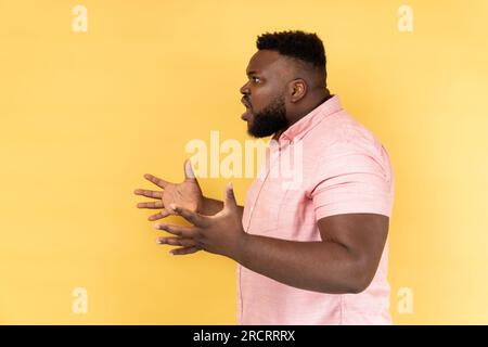 Vista laterale di un uomo infastidito e stressato che indossa una camicia rosa che ti chiede cosa vuoi e che ti spalanca le mani, litigi familiari, malintesi. Riprese in studio in interni isolate su sfondo giallo. Foto Stock