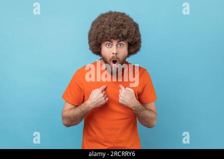 Ritratto di un uomo con acconciatura afro in maglietta arancione punta se stesso, con un'espressione sorpresa, scioccato e fiero della vittoria, incredibile successo. Riprese in studio in interni isolate su sfondo blu. Foto Stock