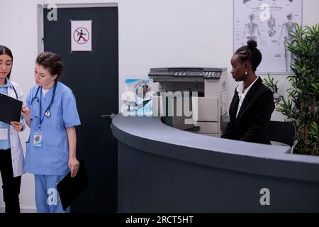 Receptionist afro-americano in piedi al banco della reception che controlla la consultazione medica sul computer durante la visita di controllo in ospedale. Lavoratore che pianifica gli appuntamenti di follow-up per i pazienti. Foto Stock