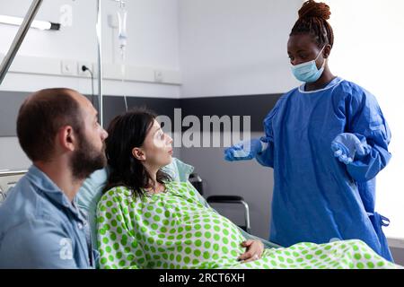 Chirurgo afroamericano che prepara la donna incinta per l'intervento chirurgico, spiegando il processo di parto nel reparto ospedaliero. Paziente con gravidanza sdraiato a letto con contrazioni che entrano in travaglio Foto Stock