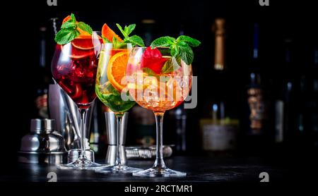 Cocktail alcolici estivi con sangria rosa, rosso, bianco, bevande con vino spagnolo, frutta, agrumi e ghiaccio. Sfondo nero del bancone del bar, utensili da bar in acciaio e. Foto Stock