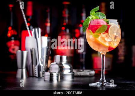 Sangria rosa bevanda alcolica estiva con vino rosa, pesca, arancia, lime e ghiaccio. Sfondo nero bar, utensili da bar in acciaio e bottiglie Foto Stock