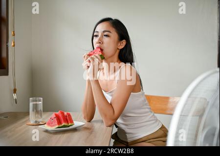 Una bella donna asiatica che sente disagio dal clima caldo, mangia anguria e siede di fronte a un ventilatore elettrico per rinfrescarsi a casa Foto Stock