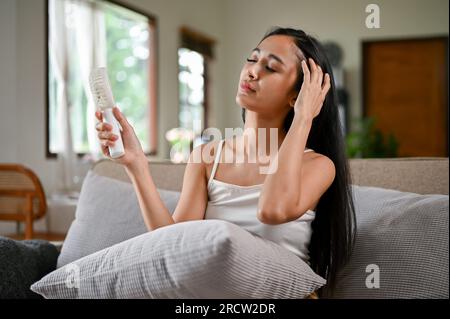 Una donna asiatica surriscaldata che sente disagio da un attacco di calore, utilizza un pratico ventilatore elettrico per raffreddarsi mentre si riposa su un divano nella sua vita Foto Stock