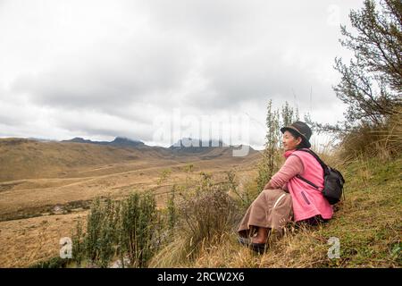 Una donna indigena solitaria seduta sulla cima di una collina si affaccia sul selvaggio e desolato alto paese delle Ande vicino a Quito, in Ecuador. Foto Stock