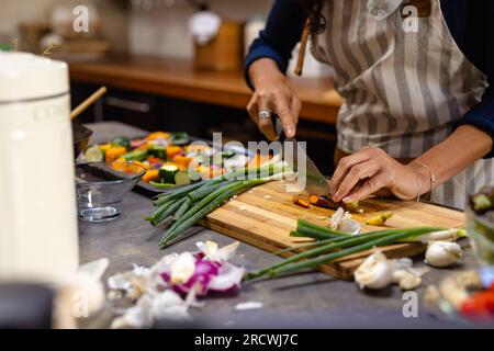 Parte centrale della donna birazziale che indossa la cena di cucina del grembiule e trita le verdure in cucina Foto Stock