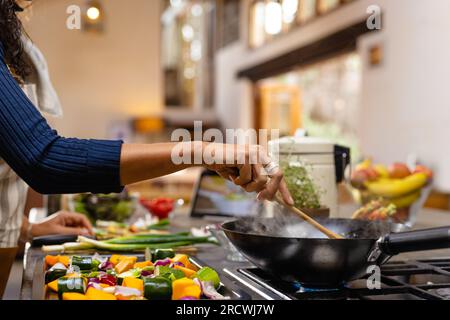 Sezione centrale della donna birazziale che indossa il grembiule che cucina la cena in cucina Foto Stock