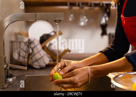 Sezione centrale di donna birazziale che indossa grembiule che lavano limone in cucina Foto Stock