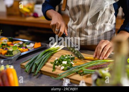 Parte centrale della donna birazziale che indossa la cena di cucina del grembiule e trita le verdure in cucina Foto Stock
