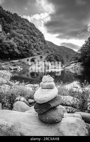 Piccola pila Zen di pietre, vista poco profonda di fronte al ponte del Diavolo sul fiume Arda, Bulgaria meridionale. Immagine di viaggio in bianco e nero Foto Stock