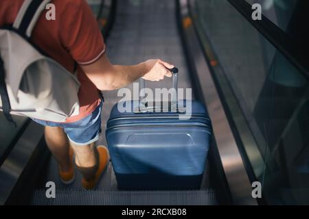 Viaggiatore che attraversa a piedi il terminal dell'aeroporto. Vista posteriore dell'uomo con valigia e zaino in piedi sull'ecalatore. Il passeggero è pronto per il viaggio. Foto Stock
