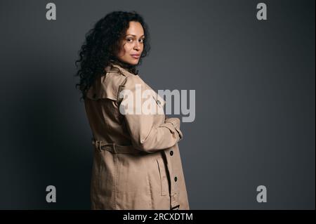 Ritratto in studio sicuro di una bella donna incinta dai capelli ricci latinoamericani, che indossa un elegante trench beige e guarda la fotocamera attraverso di lei Foto Stock