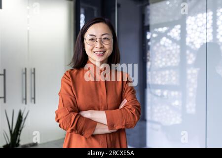 Ritratto di una giovane programmatrice asiatica di successo e sicura di sé, sviluppatrice ed esperta di IT. Si trova nel centro dell'ufficio con le braccia incrociate sul petto e guarda la telecamera con un sorriso. Foto Stock