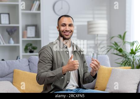 Giovane ispanico che posa per la fotocamera tenendo un bicchiere di acqua pulita e mostrando un super dito. Uomo sorridente seduto sul divano a casa. Foto Stock