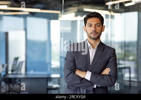 Ritratto di un uomo d'affari maturo e serio, arabo con le braccia incrociate che pensa alla fotocamera, capo in tuta da lavoro all'interno dell'ufficio in piedi vicino alla finestra. Foto Stock
