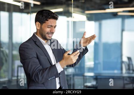 Uomo turbato e deluso che legge cattive notizie online utilizzando l'app sullo smartphone, uomo d'affari depresso sul posto di lavoro in piedi vicino alla finestra all'interno dell'ufficio con il telefono, capo arabo in tuta da lavoro. Foto Stock