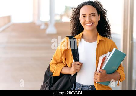 Primo piano di una graziosa studentessa brasiliana o ispanica dai capelli ricci, con uno zaino, tenere libri e quaderni in mano, stare vicino al campus universitario, guardare e sorridere alla fotocamera Foto Stock