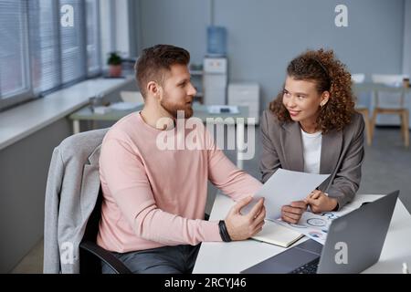 Giovane broker di sesso maschile sicuro di sé con un documento finanziario che spiega qualcosa alla collega donna sorridente durante la relazione durante la riunione di lavoro in ufficio Foto Stock