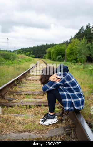 Stress, tristezza, solitudine, dolore. Una ragazza sconvolta siede sulle rotaie, una situazione psicologica, aiuto e supporto. Foto verticale Foto Stock