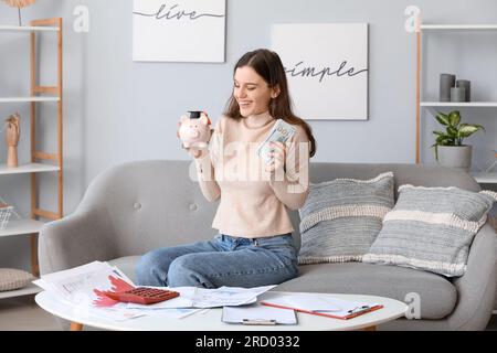 Studentessa felice che tiene salvadanaio e banconote da dollaro in soggiorno. Concetto di prestito studentesco Foto Stock
