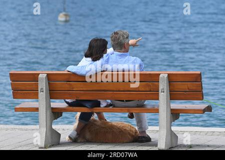 Rottach Egern, Deutschland. 17 luglio 2023. Una coppia anziana con un cane siede su una panchina a Rottach Egern sulle rive del lago Tegernsee il 07/17/2023 l'uomo mette il braccio intorno alla moglie? Credito: dpa/Alamy Live News Foto Stock