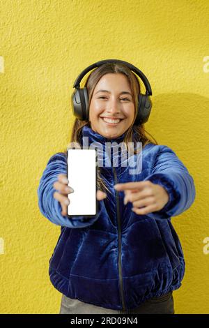 La ragazza latina con le cuffie mostra lo schermo vuoto del suo telefono cellulare isolato su uno sfondo giallo. Foto Stock