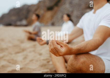 Gruppo di giovani irriconoscibili che meditano insieme, seduti in posizione di loto, praticano yoga sulla spiaggia, primo piano Foto Stock