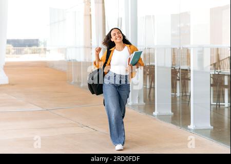 Foto a lunghezza intera di una studentessa di razza mista allegra e sorprendente con i capelli ricci, con uno zaino, tenere libri, quaderni in mano, camminare vicino all'università, guardare lontano e sorridere, finito la giornata scolastica Foto Stock