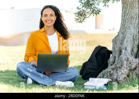 Foto a lunghezza intera di una bella ed elegante studentessa brasiliana o ispanica, seduta sull'erba vicino all'albero, con un computer portatile e un blocco note, che fa i compiti, prende appunti, sorride alla fotocamera Foto Stock