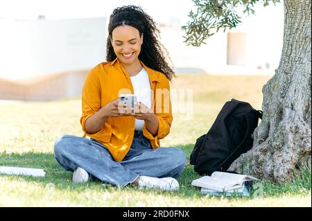 Foto a lunghezza intera di una bella ed elegante studentessa brasiliana o ispanica, seduta sull'erba vicino all'albero, usando il cellulare, scrivendo sui social media con gli amici, sorridendo felicemente Foto Stock