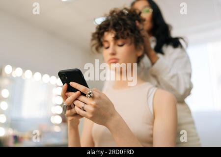 Vista ravvicinata da un'angolazione ridotta di una giovane cliente seduto su una sedia nel salone di bellezza utilizzando il telefono mentre il maestro parrucchiere offre un servizio di salone di bellezza e. Foto Stock