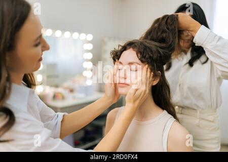 Foto media di truccatore professionista che applica il correttore sul viso di una giovane donna usando le mani per un trucco perfetto, facendo un massaggio. Foto Stock