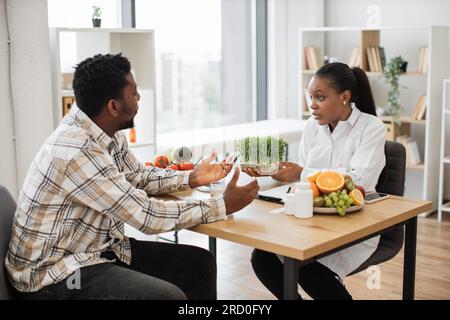 Specialista medico sicuro che mostra contenitore con microgreen all'uomo adulto in abiti casual seduti alla scrivania dell'ufficio. Professione nutrizionale multiculturale Foto Stock