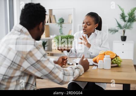 Specialista medico sicuro che mostra contenitore con microgreen all'uomo adulto in abiti casual seduti alla scrivania dell'ufficio. Nutrizione multiculturale professionale che propone una buona aggiunta a una dieta sana. Foto Stock