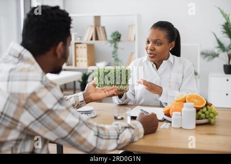 Specialista medico sicuro che mostra contenitore con microgreen all'uomo adulto in abiti casual seduti alla scrivania dell'ufficio. Professione nutrizionale multiculturale Foto Stock