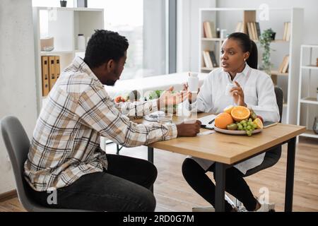 Dietista efficiente con una bottiglia di pillole in mano che parla con un uomo multirazziale seduto alla scrivania nello studio medico. Specialista medico che dà consigli su integratori vitaminici per un'alimentazione sana. Foto Stock