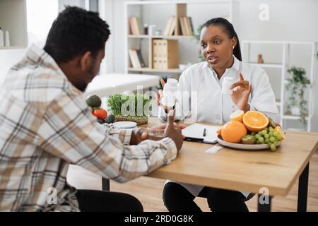 Dietista efficiente con una bottiglia di pillole in mano che parla con un uomo multirazziale seduto alla scrivania nello studio medico. Specialista medico che dà consigli su integratori vitaminici per un'alimentazione sana. Foto Stock