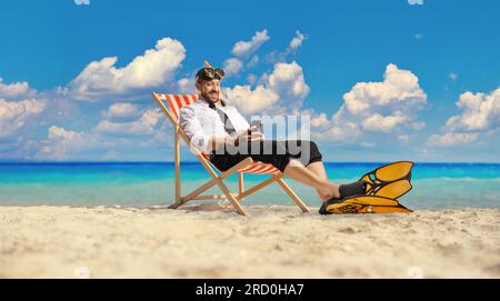 Uomo d'affari con pinne da snorkeling e maschera seduto su una sedia bech vicino al mare e digitando su uno smartphone Foto Stock