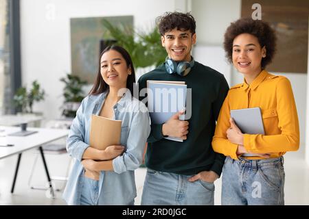 Felici tre studenti multirazziali che fingono in uno spazio di coworking, giovani con libri di lavoro in classe Foto Stock