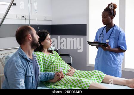 I futuri genitori discutono con l'infermiera afroamericana prima del parto nel reparto ospedaliero. Assistente medico che digita il livello di contrazioni del paziente sul computer tablet, spiegando il processo di gravidanza del travaglio Foto Stock