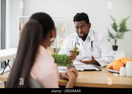 Uomo afro-americano concentrato in un cappotto bianco che tiene avocado mentre parla con una cliente femminile in una sala di consulenza. Specialista medico in cibo incoraggiando ad arricchire mangiare con frutta piena di nutrienti. Foto Stock