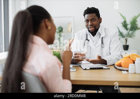 Attraente uomo barbuto in cappotto medico gesturing a vetro con fluido durante l'incontro con paziente multietnico. Nutrizionista sorridente che consiglia acqua pulita come aiuto per ridurre l'assunzione di calorie durante la dieta. Foto Stock