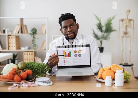 Gastroenterologo afroamericano che presenta grafici sul portatile personale e mostra il tasso di incidenza delle malattie dello stomaco a causa di un cattivo consumo. Medico barbuto in camice esortato a evitare prodotti nocivi. Foto Stock