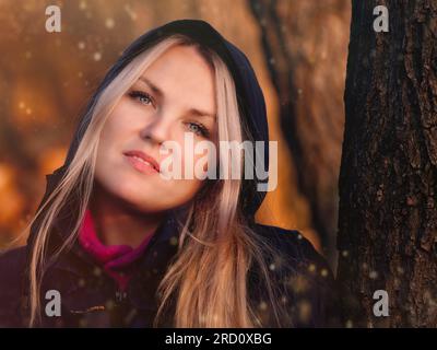 Ritratto di una giovane donna bionda in un cappuccio scuro accanto a un albero, con luce autunnale soffusa e atmosfera da sogno, un'immagine di vita all'aperto mooide Foto Stock