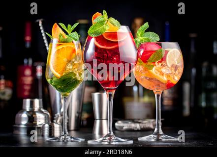 Cocktail alcolici estivi con sangria rosa, rosso, bianco, bevande con vino spagnolo, frutta, agrumi e ghiaccio. Sfondo nero del bancone del bar, utensili da bar in acciaio e. Foto Stock