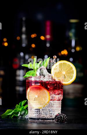 Cocktail alcolici bramble con gin secco, liquore, sciroppo, succo di limone, mora e ghiaccio. Sfondo nero bar, strumenti da bar in acciaio e bottiglia Foto Stock